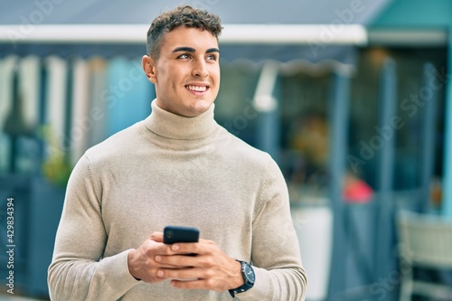 Young hispanic man smiling happy using smartphone at the city.