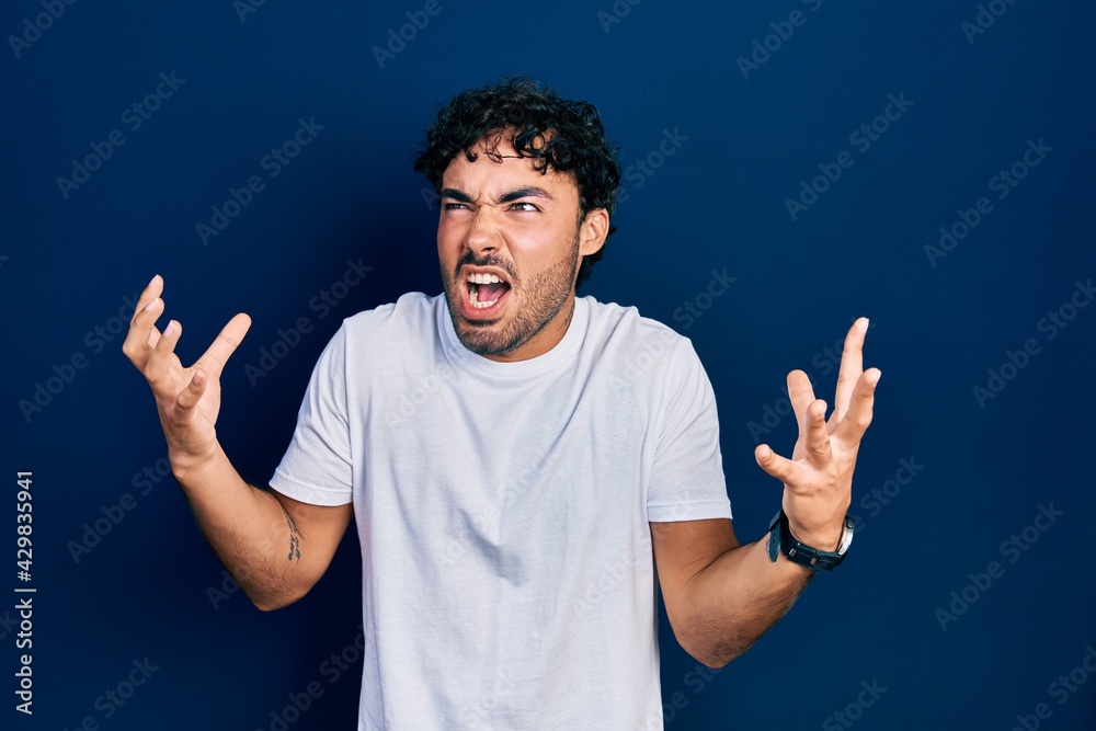 Young hispanic man wearing casual white t shirt crazy and mad shouting and yelling with aggressive expression and arms raised. frustration concept. Young hispanic man wearing casual white t shirt crazy and mad shouting and yelling with aggressive expression and arms raised. frustration concept.