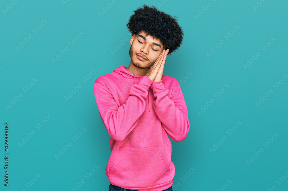 Young african american man with afro hair wearing casual pink sweatshirt sleeping tired dreaming and posing with hands together while smiling with closed eyes.