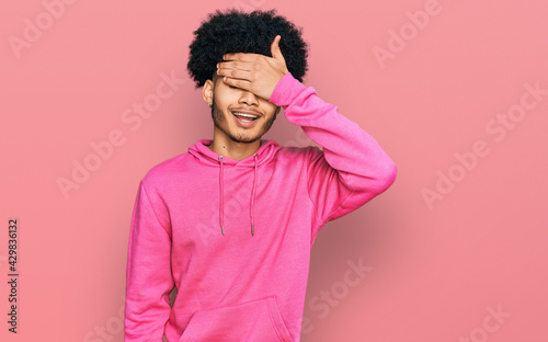 Young african american man with afro hair wearing casual pink sweatshirt smiling and laughing with hand on face covering eyes for surprise. blind concept.