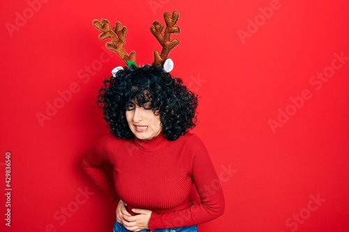 Young middle east woman wearing cute christmas reindeer horns with hand on stomach because nausea, painful disease feeling unwell. ache concept.