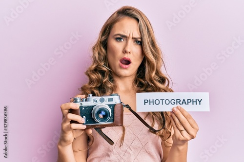 Young blonde girl holding vintage camera and paper with photography word paper in shock face, looking skeptical and sarcastic, surprised with open mouth