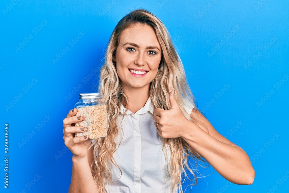 Beautiful young blonde woman holding jar with rice smiling happy and positive, thumb up doing excellent and approval sign Beautiful young blonde woman holding jar with rice smiling happy and positive, thumb up doing excellent and approval sign
