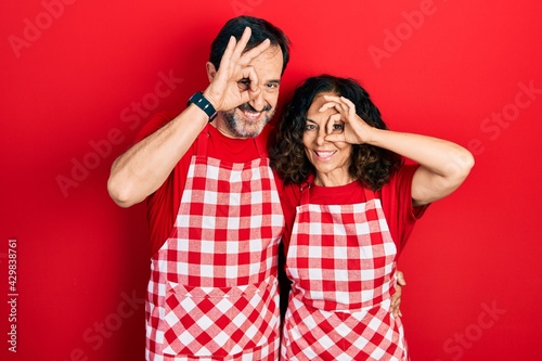 Middle age couple of hispanic woman and man wearing cook apron doing ok gesture with hand smiling, eye looking through fingers with happy face.