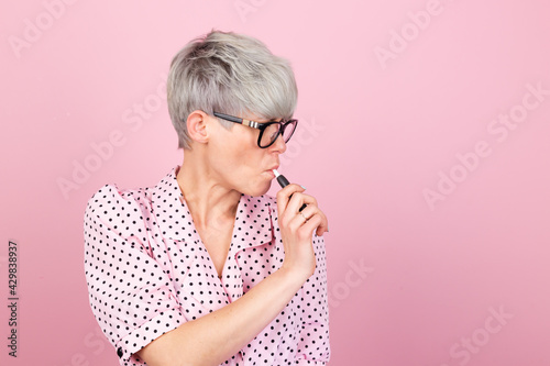 Fotografie Stylish woman in dress and glasses on pink background holding heated tobacco IQO