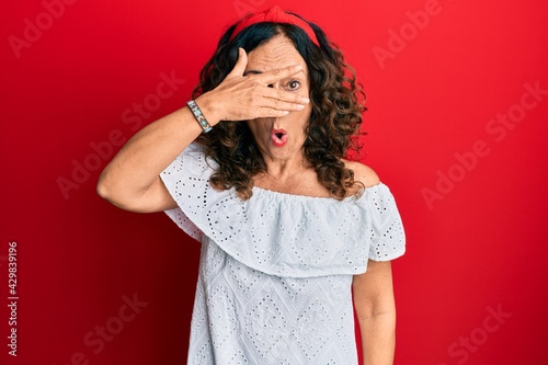 Obraz na plátně Middle age hispanic woman wearing casual clothes peeking in shock covering face and eyes with hand, looking through fingers with embarrassed expression