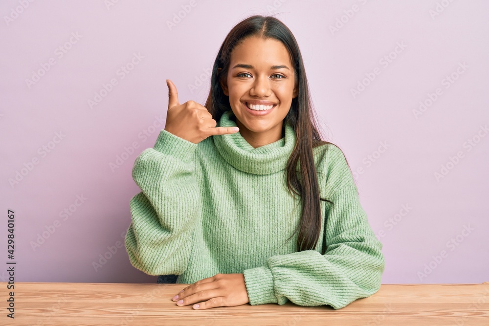 Beautiful hispanic woman wearing casual clothes sitting on the table smiling doing phone gesture with hand and fingers like talking on the telephone. communicating concepts. Beautiful hispanic woman wearing casual clothes sitting on the table smiling doing phone gesture with hand and fingers like talking on the telephone. communicating concepts.