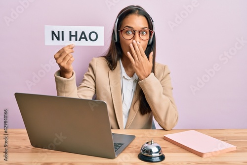 Beautiful hispanic woman wearing operator headset showing ni hao greeting covering mouth with hand, shocked and afraid for mistake. surprised expression photo