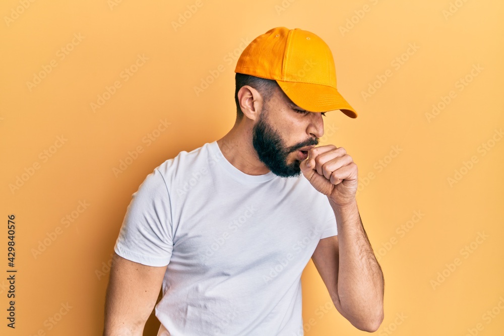 Young man with beard wearing yellow cap feeling unwell and coughing as symptom for cold or bronchitis. health care concept.