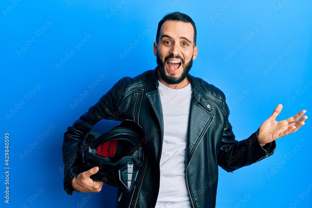 Young man with beard holding motorcycle helmet celebrating victory with happy smile and winner expression with raised hands Young man with beard holding motorcycle helmet celebrating victory with happy smile and winner expression with raised hands