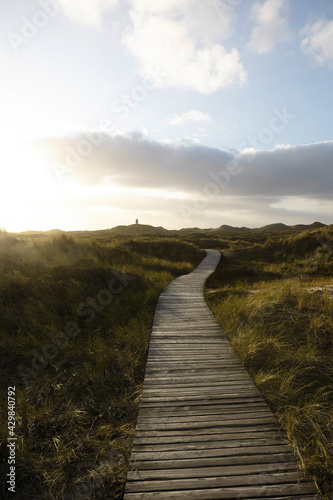 Vertically oriented photo of coastal grassy landscape. Wooden boardwalk leading to distant lighthouse building.