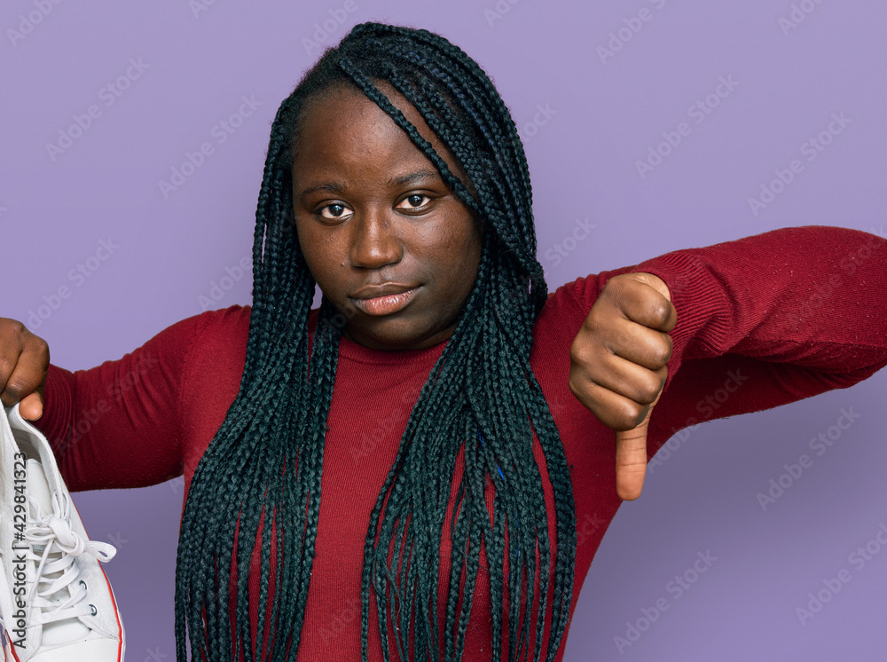 Young black woman with braids holding casual shoes with angry face, negative sign showing dislike with thumbs down, rejection concept Young black woman with braids holding casual shoes with angry face, negative sign showing dislike with thumbs down, rejection concept