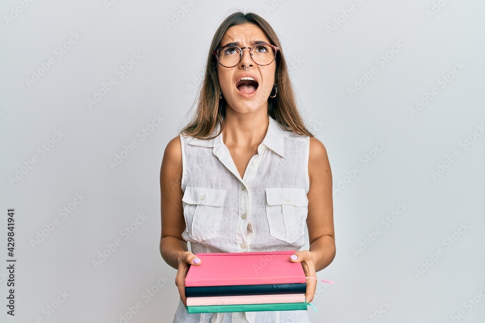 Young hispanic woman holding books angry and mad screaming frustrated and furious, shouting with anger looking up. Young hispanic woman holding books angry and mad screaming frustrated and furious, shouting with anger looking up.