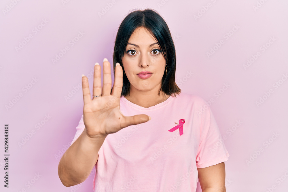Young hispanic woman wearing pink cancer ribbon on t shirt doing stop sing with palm of the hand. warning expression with negative and serious gesture on the face. Young hispanic woman wearing pink cancer ribbon on t shirt doing stop sing with palm of the hand. warning expression with negative and serious gesture on the face.