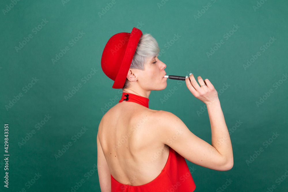 Studio portrait of stylish woman in red dress on green background with electronic cigarette IQOS heat system shows back view Studio portrait of stylish woman in red dress on green background with electronic cigarette IQOS heat system shows back view