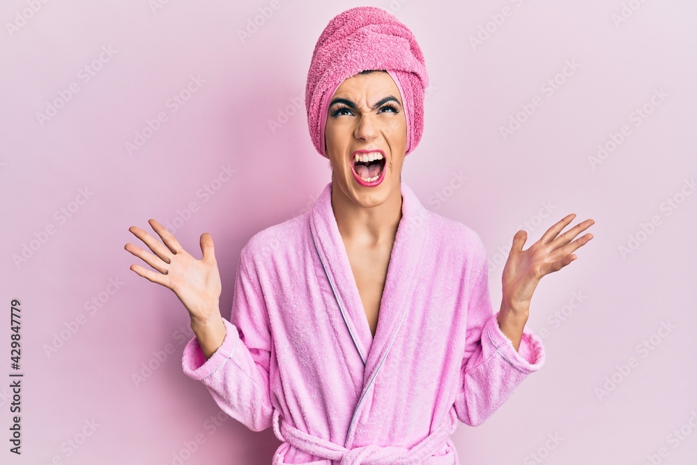 Young man wearing woman make up wearing shower towel on head and bathrobe crazy and mad shouting and yelling with aggressive expression and arms raised. frustration concept.