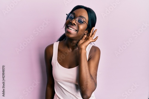Young african american woman wearing casual clothes and glasses smiling with hand over ear listening and hearing to rumor or gossip. deafness concept.