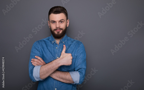 Modern happy excited young bearded man in jeans shirt shows thumbs up into the camera and having fun