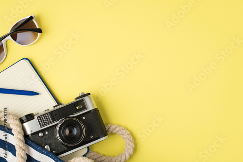 Top view photo of sunglasses beach bag with blue open diary pen and camera on isolated yellow background with copyspace