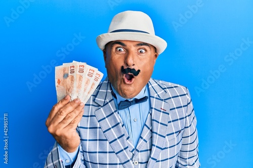 Mature middle east man with mustache wearing elegant vintage style holding pounds banknotes scared and amazed with open mouth for surprise, disbelief face photo