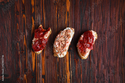 three bruschetta sandwiches on baguette bread with cottage cheese, sun-dried tomatoes and red fish on a dark wooden background, top view, copy space. Traditional italian snack for wine - grilled tapas
