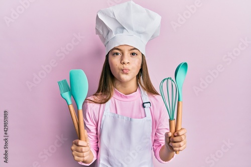 Fotografie Beautiful brunette little girl wearing professional cook apron holding cooking tools looking at the camera blowing a kiss being lovely and sexy