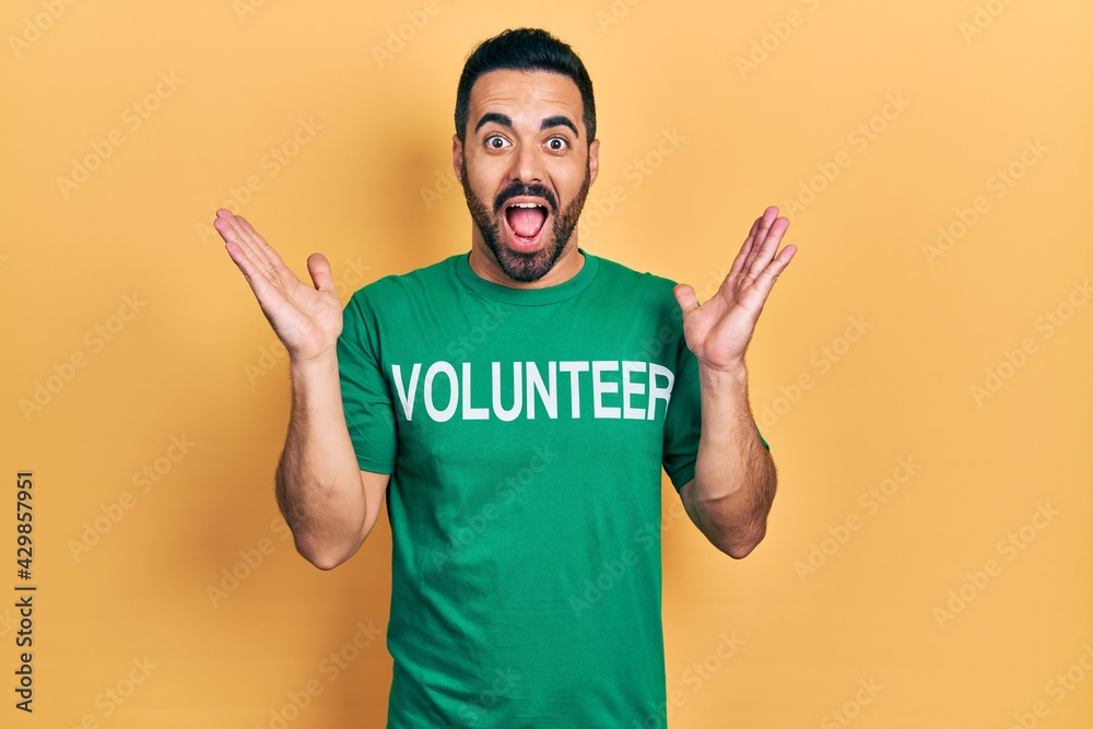 Handsome hispanic man with beard wearing volunteer t shirt celebrating victory with happy smile and winner expression with raised hands Handsome hispanic man with beard wearing volunteer t shirt celebrating victory with happy smile and winner expression with raised hands