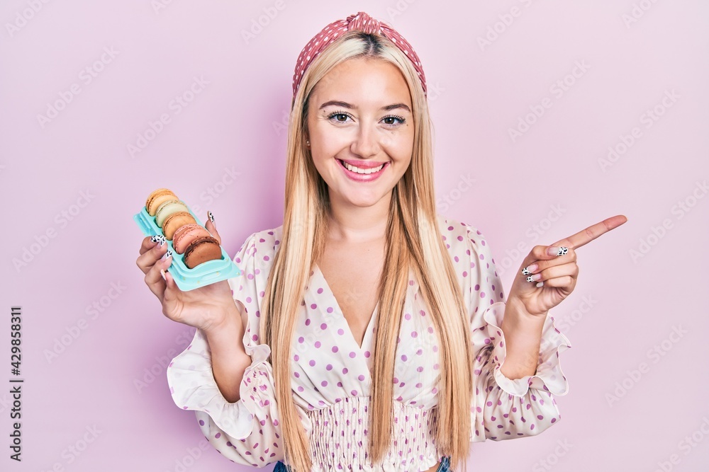 Young blonde girl holding delicious macarons pastries smiling happy pointing with hand and finger to the side Young blonde girl holding delicious macarons pastries smiling happy pointing with hand and finger to the side