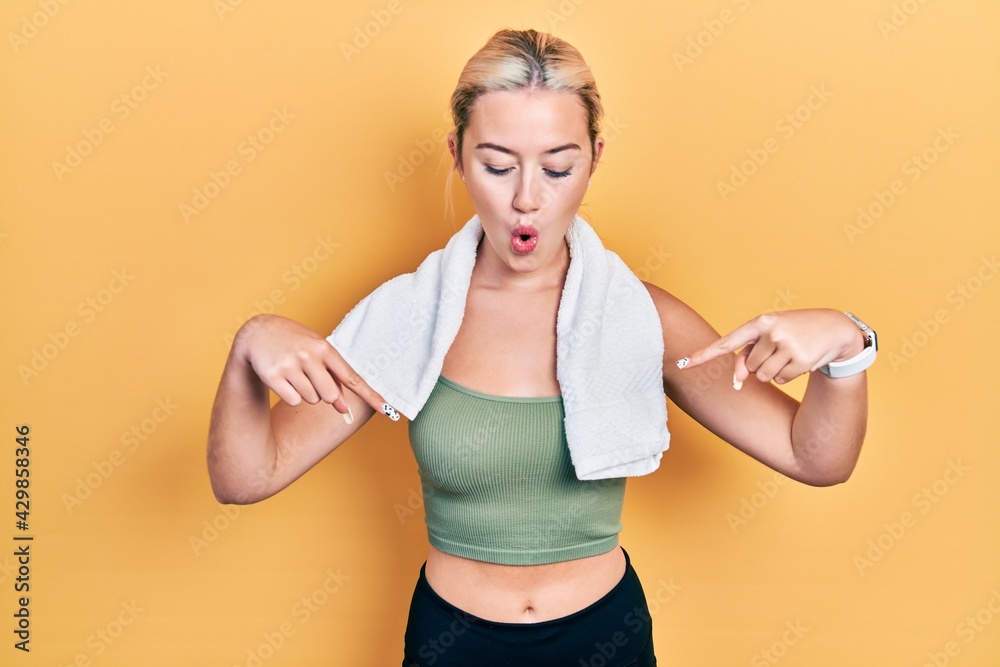 Young blonde girl wearing sportswear and towel pointing down with fingers showing advertisement, surprised face and open mouth Young blonde girl wearing sportswear and towel pointing down with fingers showing advertisement, surprised face and open mouth