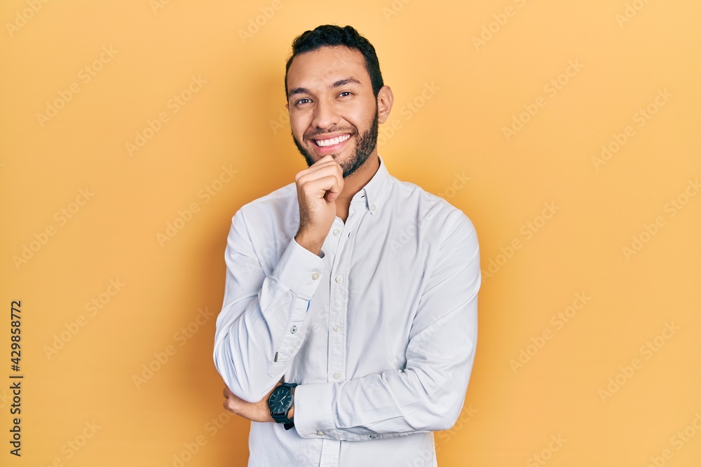 Hispanic man with beard wearing business shirt looking confident at the camera with smile with crossed arms and hand raised on chin. thinking positive. Hispanic man with beard wearing business shirt looking confident at the camera with smile with crossed arms and hand raised on chin. thinking positive.