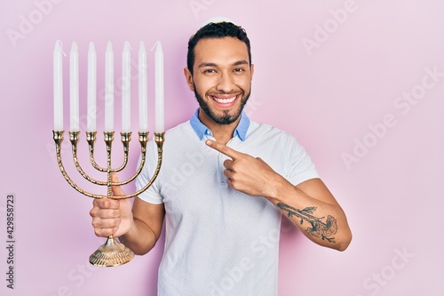 Hispanic man with beard holding menorah hanukkah jewish candle smiling happy pointing with hand and finger photo