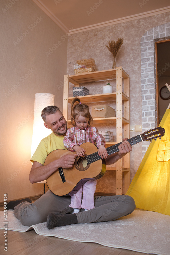 Fototapeta premium happy dad with a little daughter learns to play the guitar sitting at home on the floor near the yellow teepee