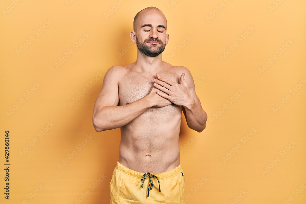 Young bald man wearing swimwear smiling with hands on chest with closed eyes and grateful gesture on face. health concept. Young bald man wearing swimwear smiling with hands on chest with closed eyes and grateful gesture on face. health concept.