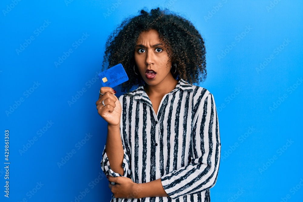 Beautiful african american woman with afro hair holding credit card in shock face, looking skeptical and sarcastic, surprised with open mouth Beautiful african american woman with afro hair holding credit card in shock face, looking skeptical and sarcastic, surprised with open mouth