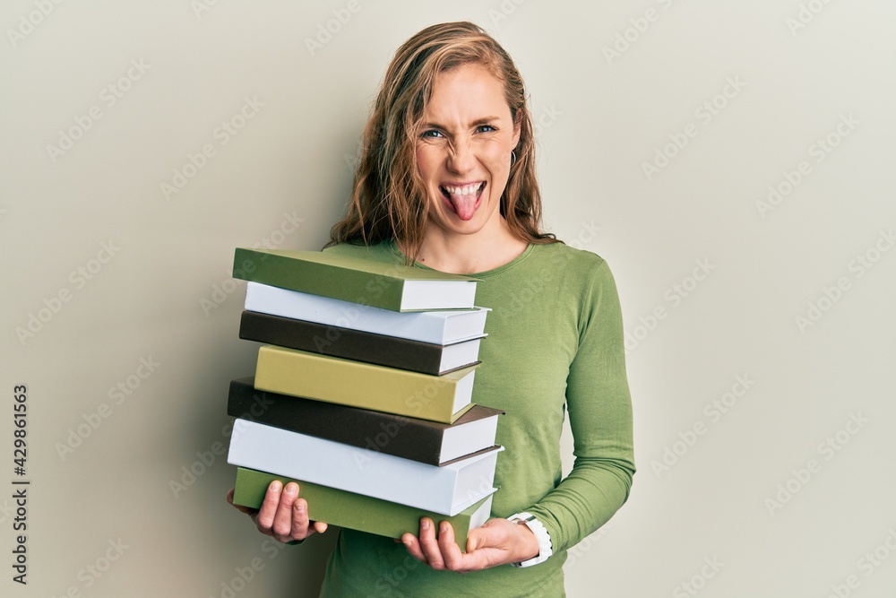 Young blonde woman holding a pile of books sticking tongue out happy with funny expression. Young blonde woman holding a pile of books sticking tongue out happy with funny expression.