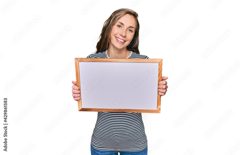 Young blonde woman holding empty white board looking positive and happy standing and smiling with a confident smile showing teeth Young blonde woman holding empty white board looking positive and happy standing and smiling with a confident smile showing teeth
