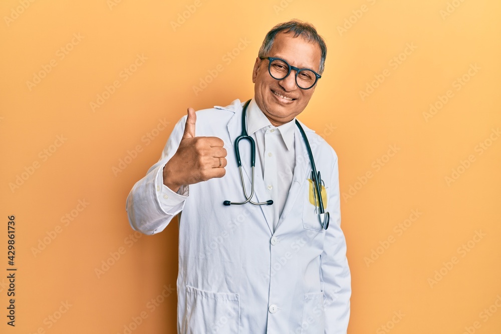 Middle age indian man wearing doctor coat and stethoscope doing happy thumbs up gesture with hand. approving expression looking at the camera showing success. Middle age indian man wearing doctor coat and stethoscope doing happy thumbs up gesture with hand. approving expression looking at the camera showing success.