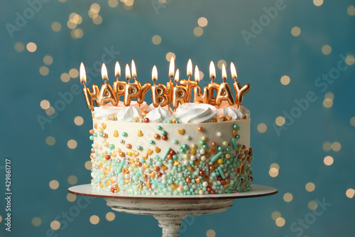 Beautiful birthday cake with burning candles on stand against festive lights