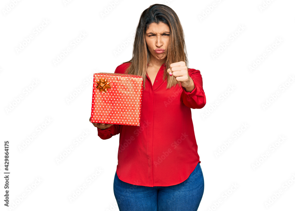 Beautiful brunette woman holding gift annoyed and frustrated shouting with anger, yelling crazy with anger and hand raised Beautiful brunette woman holding gift annoyed and frustrated shouting with anger, yelling crazy with anger and hand raised