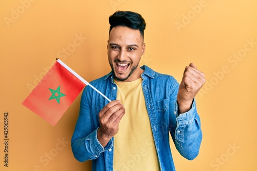 Photography Young arab man holding morocco flag screaming proud, celebrating victory and suc
