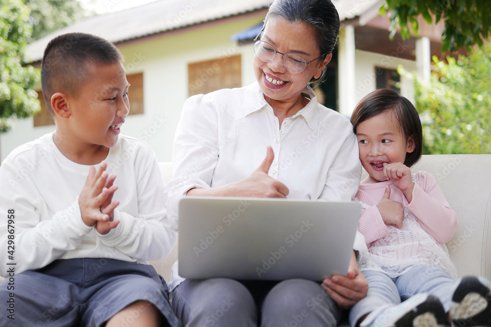 Senior Asian woman grandmother using laptop computer teaching schoolboy and little girl grandchildren, making video call with family and friends at home on house and green garden blurred background. Senior Asian woman grandmother using laptop computer teaching schoolboy and little girl grandchildren, making video call with family and friends at home on house and green garden blurred background.
