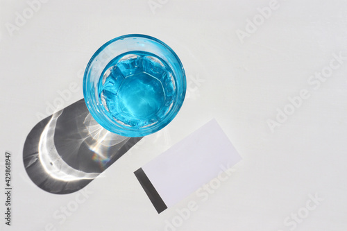 Summer still life with fresh glass of water and blank paper card on white background. Mockup invitation on white desk. Top view, copy space. Hard light, shadow
