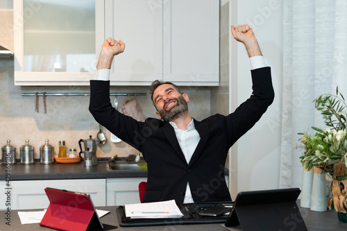 Portrait of joyful young businessman raising both arms for a deal at dawn. Expressive home worker rejoices in the kitchen. Happiness, successful meeting and contract signing.