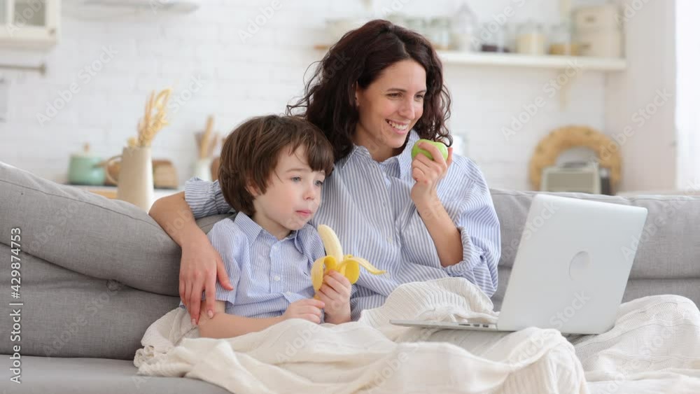 Smiling mother with little son watch funny video, cartoon on laptop computer sit together on sofa in living room. Mom and cute boy with apple and banana fruit under blanket spend time at funny movie Smiling mother with little son watch funny video, cartoon on laptop computer sit together on sofa in living room. Mom and cute boy with apple and banana fruit under blanket spend time at funny movie