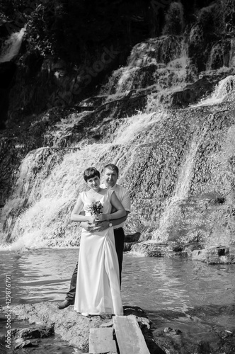 Fototapeta Naklejka Na Ścianę i Meble -  The bride and groom stand against the background of a waterfall. Newlyweds on the background of a waterfall