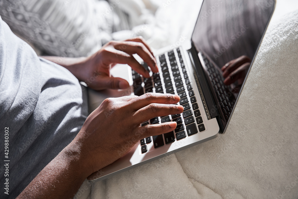 Man keeping his hands at the laptop keyboard while sitting at the bed and working