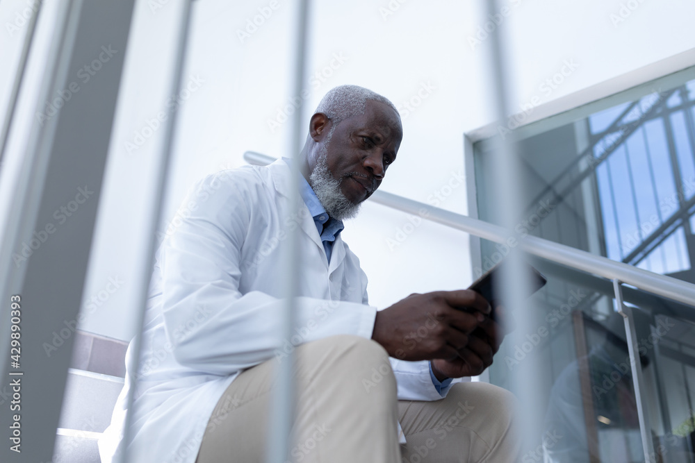 Fototapeta premium Senior african american male doctor using tablet sitting on stairs in hospital