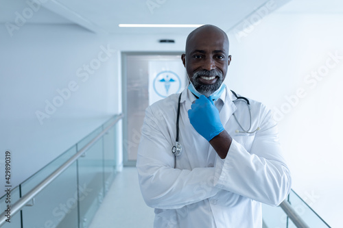 Portrait of smiling african american male doctor in hospital corridor holding face mask using tablet