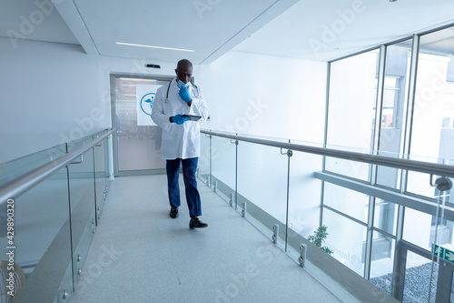 Portrait of african american male doctor walking in hospital corridor wearing face mask using tablet