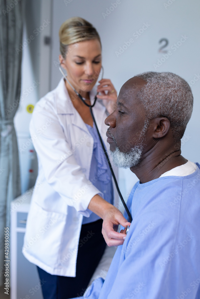 Fototapeta premium Caucasian female doctor examining with stethoscope african american male patient in hospital room